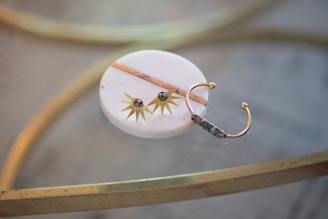 A decorative item rests on a smooth, round, white marble surface. It features a golden sunburst design with small stones at the center. A gold, open-ended band with intricate detailing sits beside it, possibly part of the overall decorative arrangement or jewelry piece. The background includes a reflective glass table and metallic gold accents.