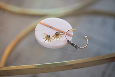A decorative item rests on a smooth, round, white marble surface. It features a golden sunburst design with small stones at the center. A gold, open-ended band with intricate detailing sits beside it, possibly part of the overall decorative arrangement or jewelry piece. The background includes a reflective glass table and metallic gold accents.