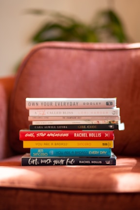 A stack of hardcover books is neatly arranged on an orange-brown armchair. The titles are visible and include topics related to self-help and personal development. The background shows a blur of greenery, suggesting a cozy indoor setting.