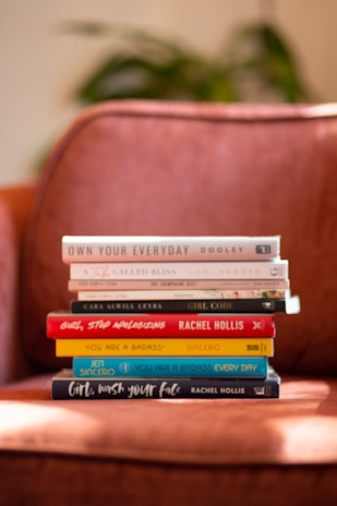 A stack of hardcover books is neatly arranged on an orange-brown armchair. The titles are visible and include topics related to self-help and personal development. The background shows a blur of greenery, suggesting a cozy indoor setting.