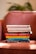 A stack of hardcover books is neatly arranged on an orange-brown armchair. The titles are visible and include topics related to self-help and personal development. The background shows a blur of greenery, suggesting a cozy indoor setting.