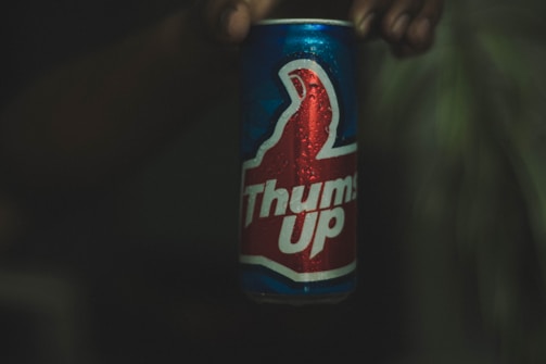 A hand holding a can of Thums Up, a carbonated drink. The can is predominantly blue with red and white highlights, featuring a thumbs up graphic. Water droplets cover the surface, suggesting coolness or condensation. The background is dark, making the can the focal point.