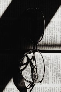 A pair of eyeglasses rests open on a page of text, with light creating dramatic shadows across the lenses and page. The shadows and light interplay focus on the detailed patterns of the text, creating a striking contrast.