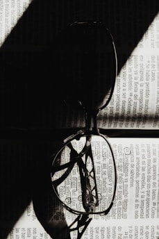 A pair of eyeglasses rests open on a page of text, with light creating dramatic shadows across the lenses and page. The shadows and light interplay focus on the detailed patterns of the text, creating a striking contrast.