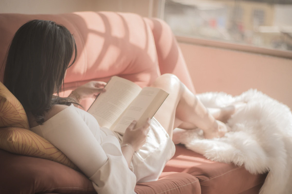 An Asian woman sitting comfortably while holding an open book, reading in soft natural light