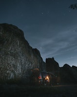 A nighttime scene with a rugged mountain or large rock formation under a starry sky. A van is parked near the rocks with its tailgate open, and a person stands illuminated by a warm light, possibly holding a lantern or flashlight.
