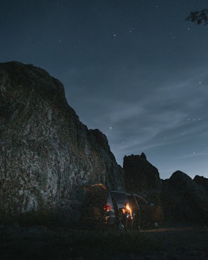 A nighttime scene with a rugged mountain or large rock formation under a starry sky. A van is parked near the rocks with its tailgate open, and a person stands illuminated by a warm light, possibly holding a lantern or flashlight.