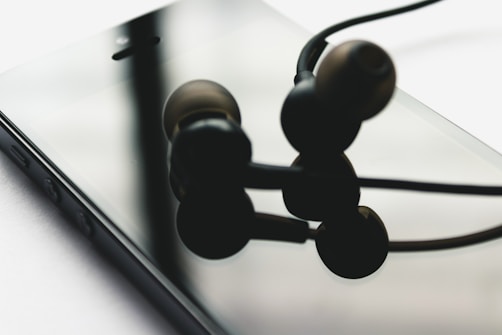 A close-up view of a smartphone lying on its screen with black in-ear headphones resting on top. The reflective surface of the phone displays a blurred reflection of the headphones.