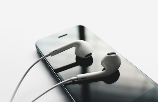 A pair of white earbuds rests on top of a black smartphone. The earbuds have a sleek, modern design and are positioned neatly against the reflective surface of the phone's screen, creating a minimalist and tech-focused composition.