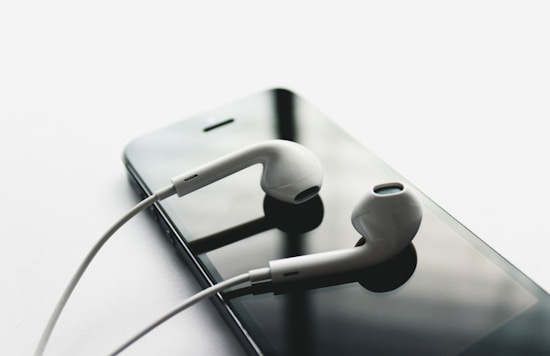 A pair of white earbuds rests on top of a black smartphone. The earbuds have a sleek, modern design and are positioned neatly against the reflective surface of the phone's screen, creating a minimalist and tech-focused composition.