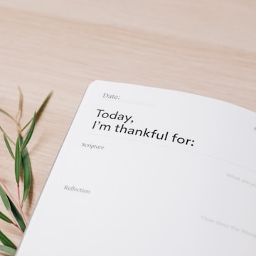 A close-up view of a journal page with headings such as 'Date' and 'Today, I'm thankful for', alongside sections titled 'Scripture' and 'Reflection'. There is also a small sprig of leaves placed on a light wooden surface.
