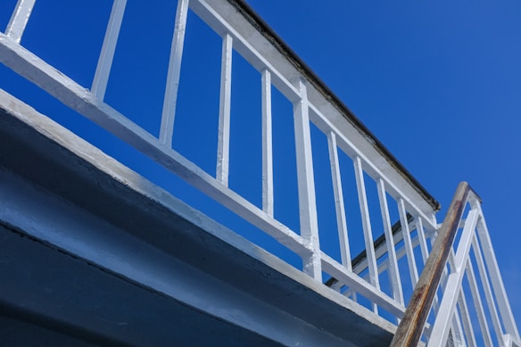 A low-angle view of a white metal staircase with railings against a clear blue sky. The staircase structure is prominently displayed with clean lines and a simple, geometric design.