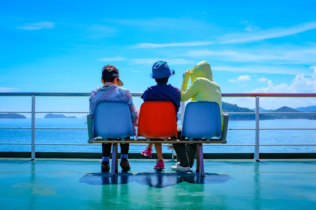 three children sitting on gang chair beside railing and ocean