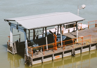 A small dock structure on a body of water with metal railings and a roof, housing boats tied to the dock. There are industrial elements such as cranes and ladders, with the dock extending into the water. The water has a muted greenish hue, and the scene is bathed in natural light.
