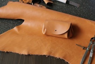 A piece of light brown leather lays across a dark speckled surface. On top of the leather is a small, neatly crafted pouch with a metallic button closure. Various crafting tools, including a compass and other leather-working instruments, are scattered around.