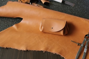 A piece of light brown leather lays across a dark speckled surface. On top of the leather is a small, neatly crafted pouch with a metallic button closure. Various crafting tools, including a compass and other leather-working instruments, are scattered around.