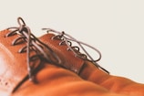 A close-up of brown leather shoes with dark laces against a light background. The focus is on the texture and stitching of the leather, highlighting the craftsmanship.