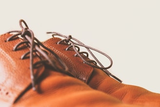 A close-up of brown leather shoes with dark laces against a light background. The focus is on the texture and stitching of the leather, highlighting the craftsmanship.
