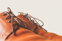A close-up of brown leather shoes with dark laces against a light background. The focus is on the texture and stitching of the leather, highlighting the craftsmanship.