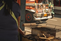 A person is standing by a street vendor stall, possibly in an urban setting. Corn and other food items are grilling on a metal barbecue grill. In the background, a colorful jeepney is passing by on the street in bright sunlight.
