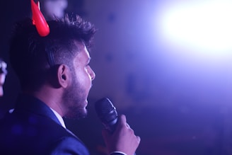 A person with short hair is holding a microphone and appears to be speaking. They are wearing red devil horns and the background is illuminated by a bright light, which creates a high contrast with the surrounding darkness.