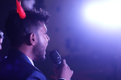 A person with short hair is holding a microphone and appears to be speaking. They are wearing red devil horns and the background is illuminated by a bright light, which creates a high contrast with the surrounding darkness.