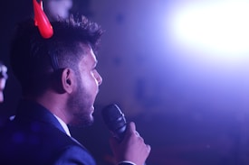 A person with short hair is holding a microphone and appears to be speaking. They are wearing red devil horns and the background is illuminated by a bright light, which creates a high contrast with the surrounding darkness.