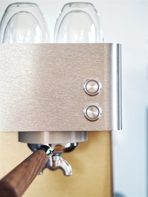 A close-up view of a stainless steel espresso machine, focusing on the stylish metallic buttons and wooden handle. Two transparent glass cups are positioned on the top of the machine.
