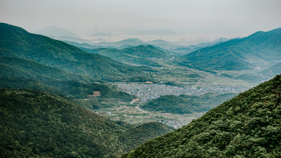 The image depicts a breathtaking view of a valley nestled between two mountains, with a lush green landscape and a clear blue sky above. The valley is dotted with small buildings scattered across its terrain, adding a touch of human presence to the natural beauty. The perspective of the photo is from a high vantage point, providing an expansive view that showcases the grandeur of the mountains and the valley below.