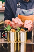 Two metallic mugs filled with a cocktail, garnished with mint leaves and dried citrus slices, are placed on a bar counter. A person wearing a dark shirt and a patterned apron is adjusting or presenting the drinks using both hands.
