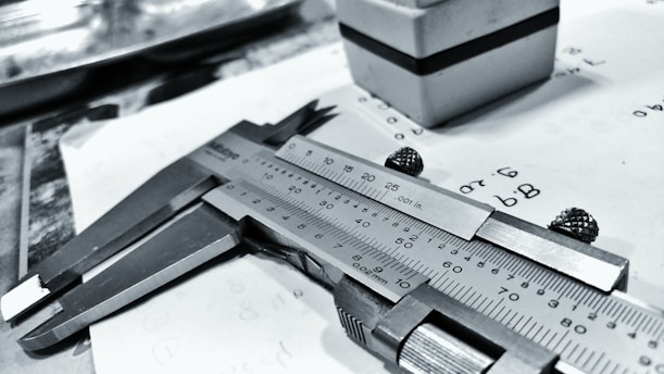 A caliper lays on a piece of paper with numerical calculations and sketches. The paper appears slightly crumpled. In the background, there is a small cuboid object with a black stripe.