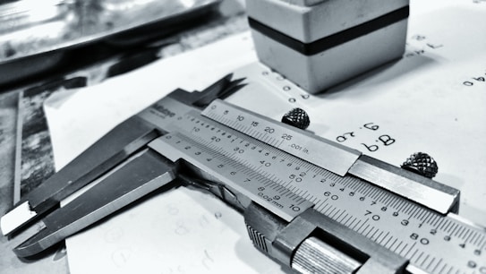 A caliper lays on a piece of paper with numerical calculations and sketches. The paper appears slightly crumpled. In the background, there is a small cuboid object with a black stripe.