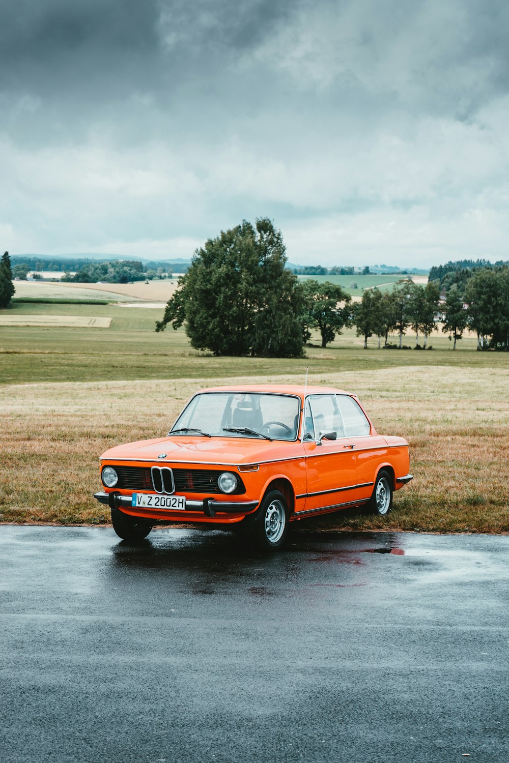 BMW coupé classica arancione parcheggiata sul ciglio della strada sotto il cielo nuvoloso grigio