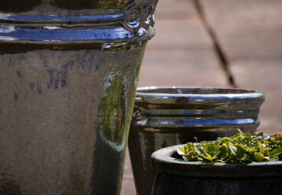 Three ceramic pots with a glossy finish featuring greenish reflections. The foreground pot is larger, while the other two are slightly smaller. One of the smaller pots has green plants growing inside. The pots are placed outdoors on a stone surface.