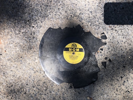 A broken vinyl record with a yellow MGM label is lying on a rough, textured concrete surface. Pieces of the record are missing, suggesting it has been shattered.