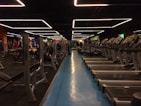 A spacious gym with multiple rows of treadmills and weightlifting benches on either side. The lighting features geometric patterns on the ceiling, creating a modern and organized ambiance. People are actively using the equipment, engaging in fitness activities.