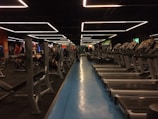 A spacious gym with multiple rows of treadmills and weightlifting benches on either side. The lighting features geometric patterns on the ceiling, creating a modern and organized ambiance. People are actively using the equipment, engaging in fitness activities.