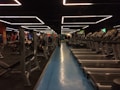 A spacious gym with multiple rows of treadmills and weightlifting benches on either side. The lighting features geometric patterns on the ceiling, creating a modern and organized ambiance. People are actively using the equipment, engaging in fitness activities.