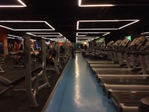 A spacious gym with multiple rows of treadmills and weightlifting benches on either side. The lighting features geometric patterns on the ceiling, creating a modern and organized ambiance. People are actively using the equipment, engaging in fitness activities.