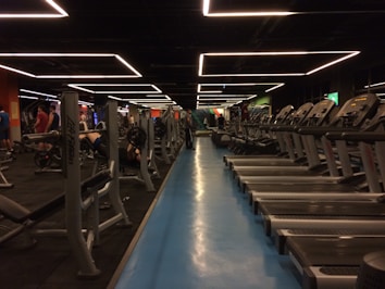 A spacious gym with multiple rows of treadmills and weightlifting benches on either side. The lighting features geometric patterns on the ceiling, creating a modern and organized ambiance. People are actively using the equipment, engaging in fitness activities.