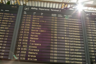An airport departure board displays a list of upcoming flights, times, destinations, and airlines, primarily featuring Lufthansa. Sunlight brightens the top corner, creating a lens flare effect.