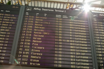 An airport departure board displays a list of upcoming flights, times, destinations, and airlines, primarily featuring Lufthansa. Sunlight brightens the top corner, creating a lens flare effect.