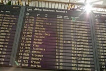 An airport departure board displays a list of upcoming flights, times, destinations, and airlines, primarily featuring Lufthansa. Sunlight brightens the top corner, creating a lens flare effect.