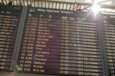 An airport departure board displays a list of upcoming flights, times, destinations, and airlines, primarily featuring Lufthansa. Sunlight brightens the top corner, creating a lens flare effect.