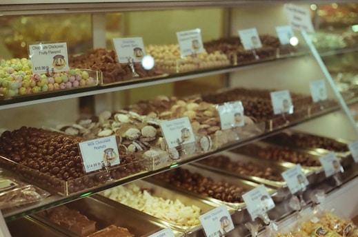 A display case filled with various types of chocolates and candies, each labeled with their name and price. The assortment includes chocolate chickpeas, chocolate peanuts, and other confections, arranged neatly on multiple shelves.