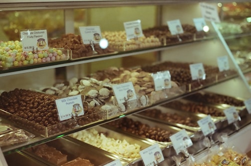 A display case filled with various types of chocolates and candies, each labeled with their name and price. The assortment includes chocolate chickpeas, chocolate peanuts, and other confections, arranged neatly on multiple shelves.