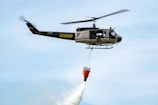 A sheriff helicopter is flying in the sky while carrying an orange water bucket designed for firefighting. The helicopter is marked with 'Lake County Sheriff' and has two people visible near the open door.
