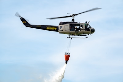 A sheriff helicopter is flying in the sky while carrying an orange water bucket designed for firefighting. The helicopter is marked with 'Lake County Sheriff' and has two people visible near the open door.