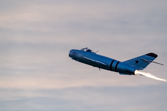 A single-engine jet aircraft with a blue-gray exterior and a star emblem on the tail flies through the sky. A trail of bright flame is visible from the engine, indicating the aircraft is in motion. The sky is cloudy with soft gray and blue hues, creating a serene and dynamic scene.