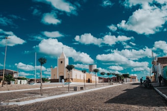 A small church with a cross on top is situated in the center of a quiet, cobblestone street lined with neatly trimmed trees. The sky is bright blue with scattered white clouds. In the background, there are a few buildings with colorful facades and a water tower.